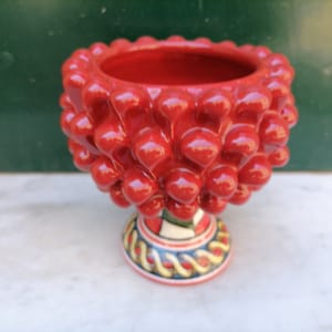 Puede incluir: Un cuenco de cerámica rojo con forma de piña con una base de patrones coloridos. El cuenco es perfecto para servir aperitivos o guardar objetos pequeños.