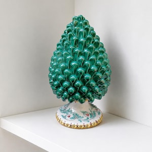 Può includere: Scultura decorativa a forma di pigna in ceramica. La pigna è di un verde vibrante con finitura lucida. Poggia su una base bianca con motivi floreali e a foglie in rosso, blu e giallo. L'oggetto è esposto su una mensola bianca.