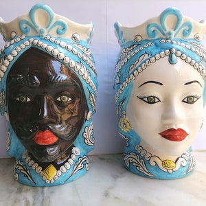 May include: Two ceramic planter vases shaped like human heads. The vase on the left has a black face with a crown and a blue and white patterned design. The vase on the right has a white face with a crown and a blue and white patterned design.