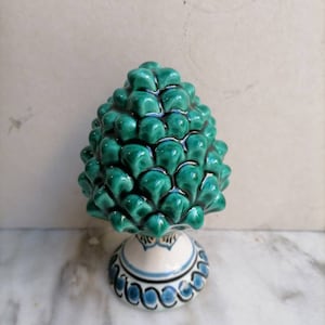 Puede incluir: Una figurita de cerámica verde en forma de piña con una base pintada de blanco y azul. La base tiene un patrón decorativo de remolinos azules y blancos.
