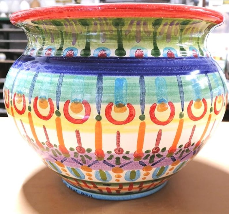 Może przedstawiać: Duży, okrągły, kolorowy ceramiczny pojemnik na rośliny z szeroką krawędzią. Pojemnik jest ozdobiony geometrycznym wzorem w odcieniach niebieskiego, zielonego, ż&oacute;łtego, pomarańczowego, czerwonego i fioletowego.