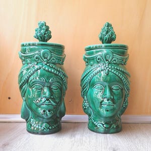 Può includere: Due vasi in ceramica verde con coperchi a forma di pigne. I vasi sono decorati con volti e dettagli intricati.