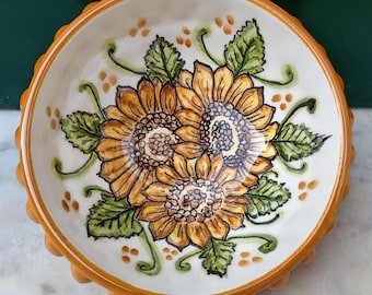 May include: A ceramic bowl with a white interior and a yellow-orange rim. The bowl features a hand-painted design of three sunflowers with green leaves and stems. The rim has a scalloped edge.