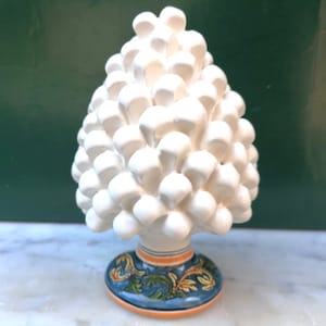 May include: White ceramic pine cone sculpture with a blue and green base with a floral design. The base has orange and yellow stripes.