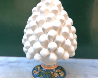 Puede incluir: Escultura de piña de cerámica blanca con una base azul y verde con un diseño floral. La base tiene rayas naranjas y amarillas.