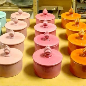 Puede incluir: Una colección de 20 tarros de cerámica con tapa en varios colores, incluyendo turquesa, azul claro, rosa, amarillo y naranja. Los tarros están dispuestos en filas sobre una superficie de madera.