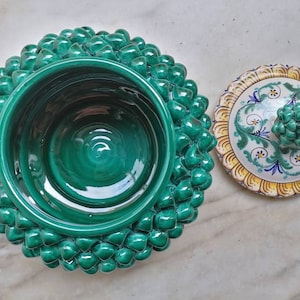 May include: A green ceramic bowl with a textured, pinecone-like exterior. The bowl has a lid with a floral design in yellow, blue, and green.