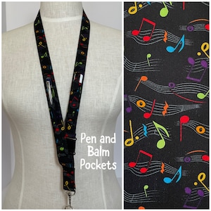 May include: Black lanyard with a repeating pattern of colorful musical notes. The lanyard has a pen pocket and a metal clasp. The text "Pen and Balm Pockets" is visible on the image.