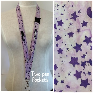 May include: A lavender lanyard with a repeating pattern of purple stars and dots. The lanyard has two pen pockets and a black plastic clasp. The text "Two pen Pockets" is printed on the lanyard.