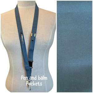 Peut inclure: Un cordon bleu avec des poches pour stylo et baume. Le cordon est fait d'un tissu robuste et comporte un fermoir noir et un clip argenté en bas. Un stylo est visible dans l'une des poches. Le texte "Pen and balm Pockets" est imprimé en bas.