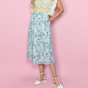 May include: A woman wearing a cream-colored lace top with ruffled sleeves and a light blue midi skirt with a floral pattern. The skirt has a button-down front and pockets. She is wearing white heels with clear block heels.