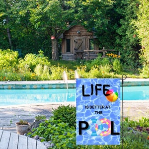 Puede incluir: Una bandera de jardín azul y blanca con el texto "Life is better at the pool" y una pelota de playa y un flotador inflable. La bandera se muestra frente a una piscina.
