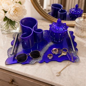 May include: A royal blue vanity tray with silver glitter accents holds a variety of items. These include a decorative container, a pen holder, sunglasses, rings, keys, and a necklace. A bouquet of white flowers is in the background.