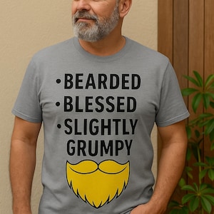 Puede incluir: Una camiseta gris con las palabras "BEARDED, BLESSED, SLIGHTLY GRUMPY" en negro y un gráfico de barba amarilla. La persona lleva vaqueros azules.