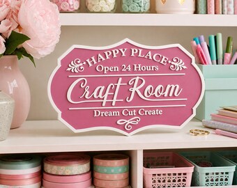 May include: A pink sign with white lettering that reads "Happy Place Craft Room Open 24 Hours Dream Cut Create." The sign is on a white shelf with jars of buttons, a vase of pink flowers, and craft supplies.