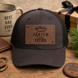 May include: A dark grey trucker cap with a black mesh back and a brown leather patch. The patch reads "World's Greatest Farter: I mean Father" in white lettering. A mug with "Best Dad Ever" is visible.