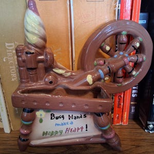 May include: A brown ceramic planter shaped like a spinning wheel with a cream-coloured top section. The front features the text "Busy Hands make a Happy Heart!" in black and blue lettering, accented by a small yellow flower graphic and colourful dots.