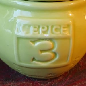 Puede incluir: Un tarro de especias de cer&aacute;mica verde claro con la palabra "SPICE" y el n&uacute;mero "3" en relieve en la parte delantera. El tarro tiene forma redondeada y un acabado brillante. El fondo es de color marr&oacute;n rojizo borroso.