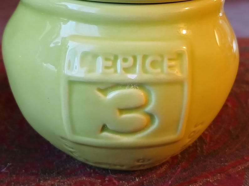 Puede incluir: Un tarro de especias de cer&aacute;mica verde claro con la palabra "SPICE" y el n&uacute;mero "3" en relieve en la parte delantera. El tarro tiene forma redondeada y un acabado brillante. El fondo es de color marr&oacute;n rojizo borroso.