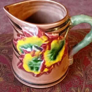 Può includere: Un'anfora in ceramica marrone con un manico verde. L'anfora è decorata con un motivo floreale di foglie rosse, gialle e verdi.