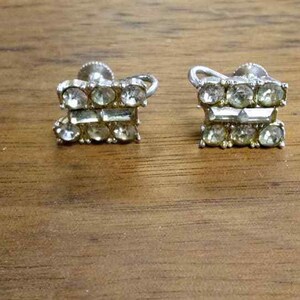 May include: A pair of silver clip-on earrings with clear rhinestones. The earrings are in the shape of a rectangle and have two rows of rhinestones.