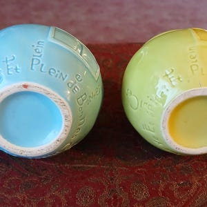 Puede incluir: Dos jarrones esf&eacute;ricos de cer&aacute;mica, uno azul claro y otro amarillo verdoso. Ambos tienen una abertura circular y texto franc&eacute;s en relieve. Los jarrones est&aacute;n sobre una superficie estampada de color marr&oacute;n rojizo.