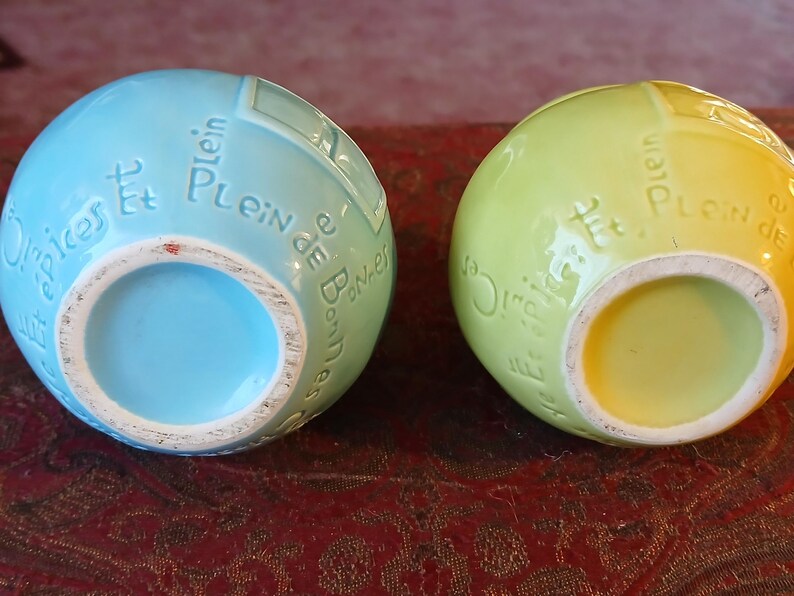 Puede incluir: Dos jarrones esf&eacute;ricos de cer&aacute;mica, uno azul claro y otro amarillo verdoso. Ambos tienen una abertura circular y texto franc&eacute;s en relieve. Los jarrones est&aacute;n sobre una superficie estampada de color marr&oacute;n rojizo.