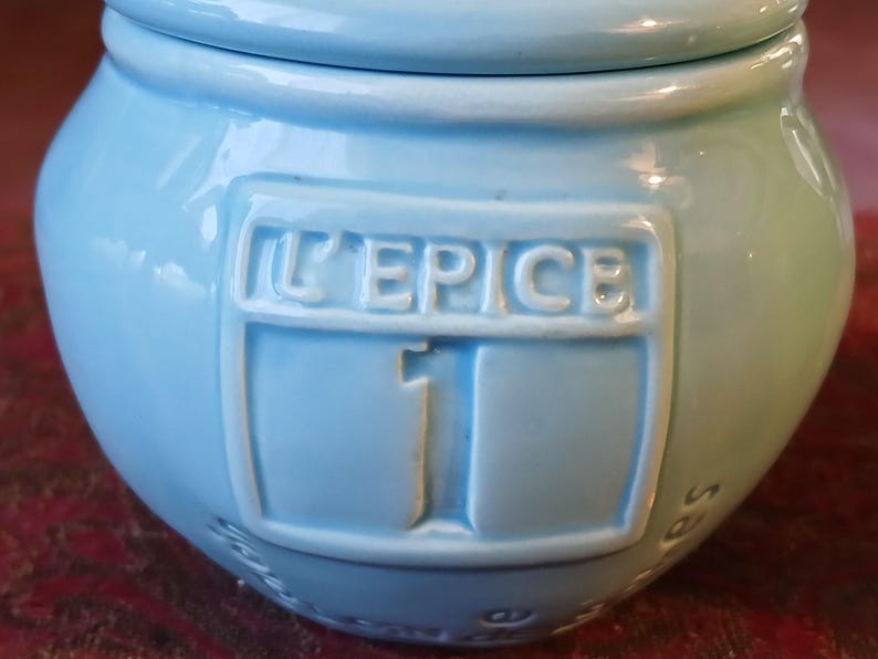 Puede incluir: Un tarro de especias de cer&aacute;mica azul claro con tapa. El tarro tiene forma redondeada y presenta el texto en relieve "L'EPICE" y el n&uacute;mero "1" en la parte delantera. El tarro se utiliza probablemente para guardar especias.