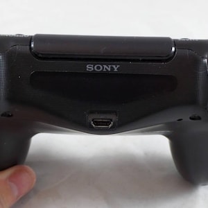 May include: A black PlayStation 4 controller with the word "SONY" printed on the front. The controller has buttons labeled "R1", "R2", "L1", and "L2".