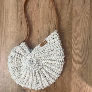 May include: A cream-colored, crocheted handbag with a brown leather strap. The bag has a circular, fan-like design and a small, rectangular tag. The strap is attached to the bag at two points.