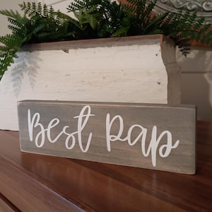 May include: A gray wooden sign with the words "best paps" written in white paint.