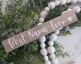 May include: A wooden sign with the text "Best Nana Ever" in white lettering and a small white heart. The sign is sitting on a bed of green foliage and white beads.