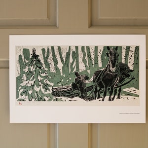 May include: A framed print showing a winter landscape with a horse-drawn sleigh transporting a log through a snowy forest. The artwork uses green and black tones, featuring detailed illustrations of trees and a figure guiding the horses.
