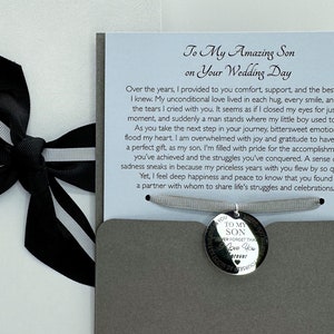 May include: A gray card with a white card inside. The white card has a black ribbon tied around it. The gray card has a message that reads "To My Amazing Son on Your Wedding Day" and a silver charm with the words "To My Son Never Forget That I Love You Always" on it.