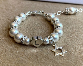 May include: A white pearl bracelet with a silver charm in the shape of a heart with the letter L on it. The bracelet also has a silver charm in the shape of a Star of David.