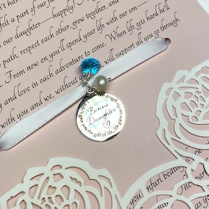 May include: A white satin ribbon with a silver charm that reads "Bonus Daughter" and features a blue gemstone and a pearl. The ribbon is tied around a pink card with white cut-out roses and the text "From now on you'll spend your life with our son and love in each adventure to come. When life gets hard he'll with you and we, without question, will support all you go through."