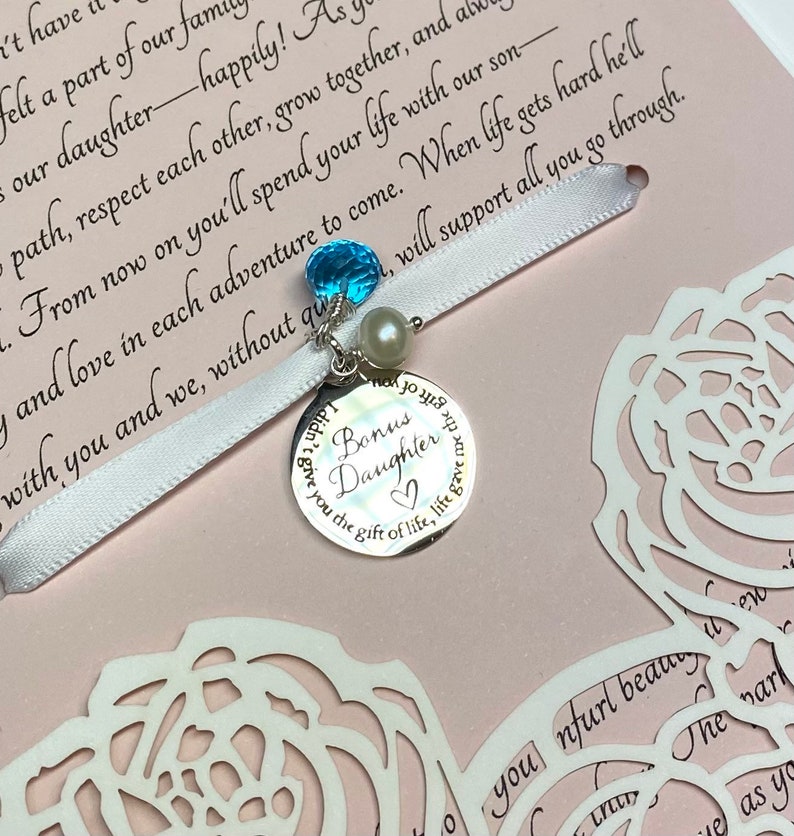 May include: A white satin ribbon with a silver charm that reads "Bonus Daughter" and features a blue gemstone and a pearl. The ribbon is tied around a pink card with white cut-out roses and the text "From now on you'll spend your life with our son and love in each adventure to come. When life gets hard he'll with you and we, without question, will support all you go through."