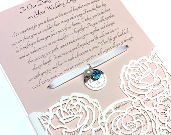 May include: A white card with a pink background and a white lace design. The card has a message to a daughter-in-law on her wedding day. A white ribbon with a pearl and blue charm is attached to the card.