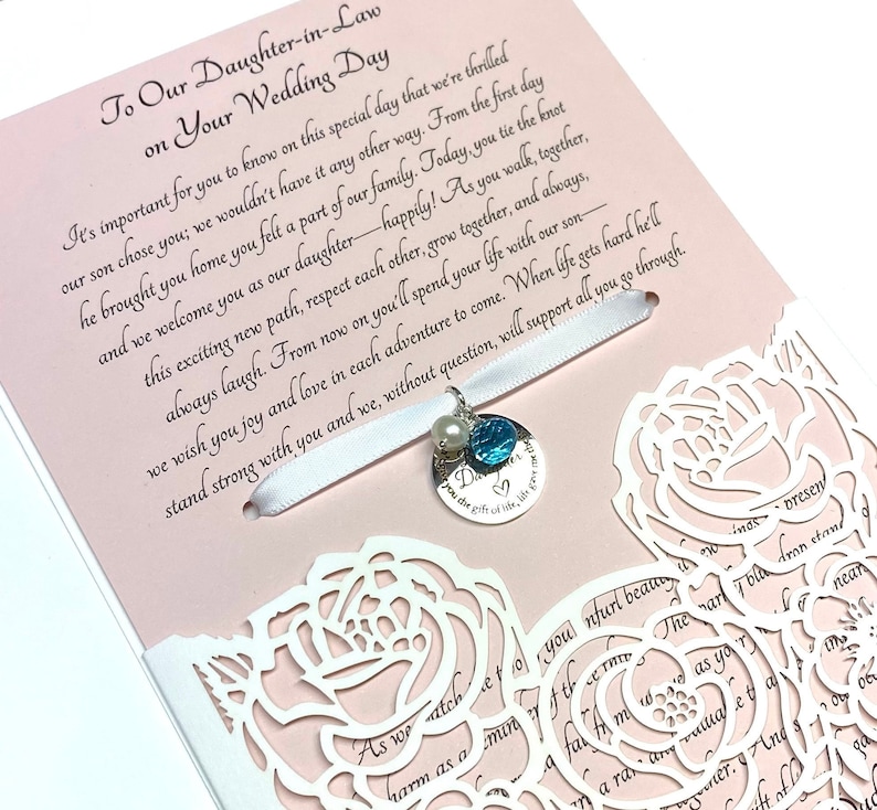 May include: A white card with a pink background and a white lace design. The card has a message to a daughter-in-law on her wedding day. A white ribbon with a pearl and blue charm is attached to the card.