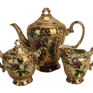 May include: A gold and white porcelain tea set with floral designs. The set includes a teapot, creamer, and sugar bowl.