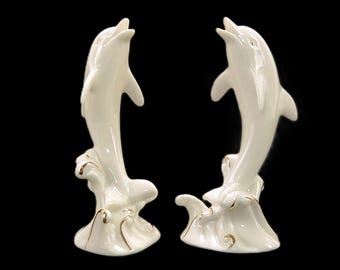 May include: Two white porcelain dolphin figurines leaping out of the water. The dolphins are facing each other and have gold accents on their fins and tails.