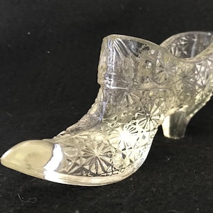 Può includere: Oggetto decorativo a forma di scarpa in vetro trasparente. La scarpa ha una punta affilata e un piccolo tacco. Il vetro è strutturato con un motivo a stella. È probabilmente un oggetto decorativo o un piccolo contenitore.