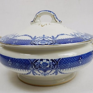 Peut inclure: Une soupière en céramique blanche avec couvercle, ornée d'un motif floral et géométrique bleu sur le corps et le couvercle. La soupière possède deux anses latérales et une poignée centrale sur le couvercle, avec des bordures dorées.