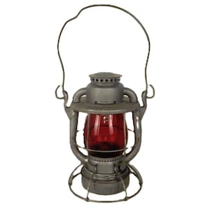 Puede incluir: Una linterna vintage de metal gris con un globo de cristal rojo. La linterna tiene un asa de alambre y una base circular. El diseño recuerda a las linternas antiguas de ferrocarril o de camping.