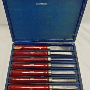 Könnte beinhalten: Ein Set aus acht Holzbeiteln mit dunkelroten Holzgriffen und silbernen Metallklingen, angeordnet in einer blauen Holzaufbewahrungsbox. Der Innendeckel der Box zeigt das Wort CRAFTSMAN in weißer Schrift auf einem blauen Hintergrund.
