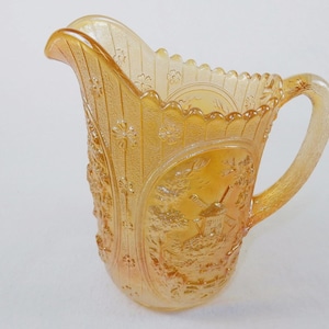 May include: An iridescent marigold carnival glass pitcher featuring a relief pattern of a windmill in a landscape scene. The pitcher has a scalloped rim, a curved handle, and vertical panels decorated with small embossed flower motifs.