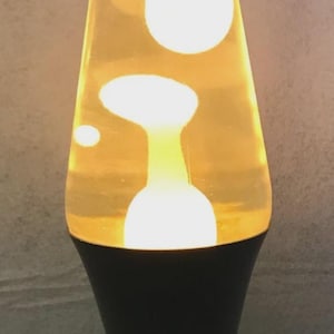 Könnte beinhalten: Eine Retro-Lavalampe mit einer transparenten Glasflasche, gefüllt mit gelber Flüssigkeit und weißen Formen. Die Lampe hat einen schwarzen Sockel und einen Metallverschluss. Die Lampe ist eingeschaltet, die Formen leuchten.