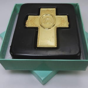May include: A gold cross with intricate patterns, displayed in a black box, which is inside a pale green gift box. The cross has a central emblem and decorative borders, possibly for religious or commemorative purposes.