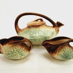 May include: A ceramic tea set consisting of a teapot with a lid, a creamer, and a sugar bowl. Each piece features a brown and cream gradient glaze with a raised, light green floral relief pattern and integrated handles with a curved design.