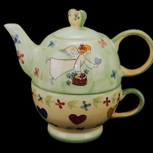 May include: A green and yellow ceramic teapot with a painted angel design. The teapot has a removable lid and a matching cup.